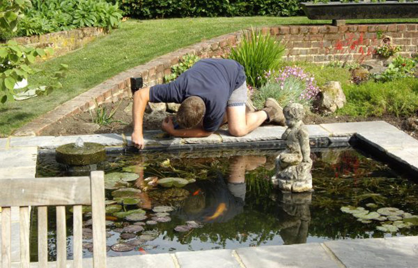 A formal sunken pond containing fish, water lilies and sculptures. A landscaper is looking into the pond adjusting the pond liner