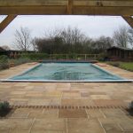 Outdoor swimming pool with surrounding paving, a timber summer house in the background and a wooden pergola in the foreground