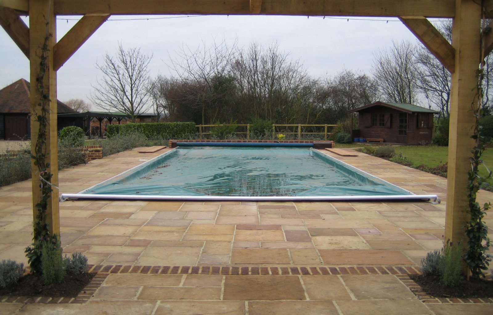 Outdoor swimming pool with surrounding paving, a timber summer house in the background and a wooden pergola in the foreground