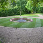 A formal round pond with fountain within turfed circular area with block edging and shingle pathway.