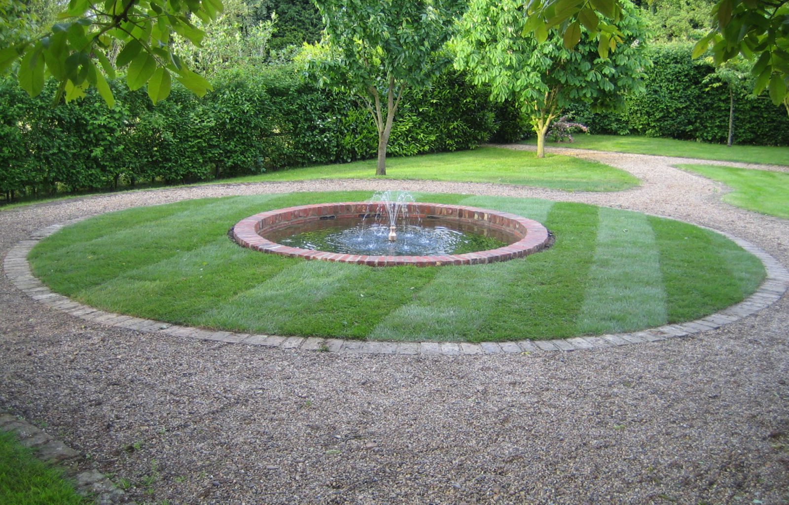 A formal round pond with fountain within turfed circular area with block edging and shingle pathway.