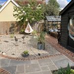 A domestic garden that features paved patio, stone covered borders, planted containers, a green house, hammock and a shed.