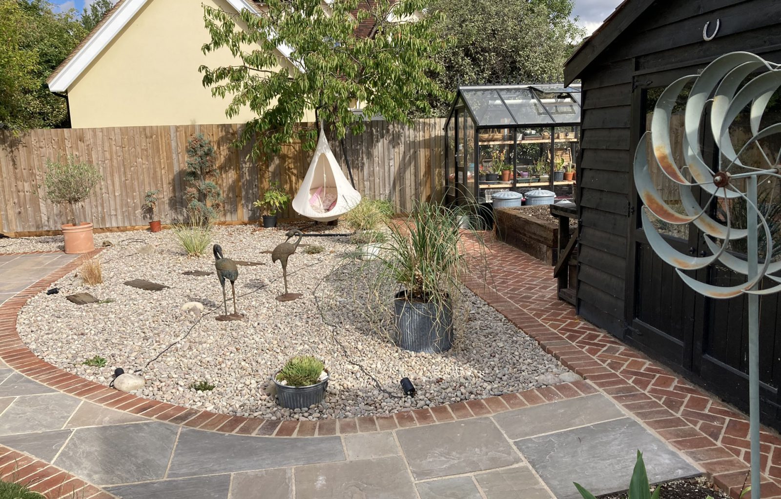 A domestic garden that features paved patio, stone covered borders, planted containers, a green house, hammock and a shed.