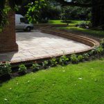 A large sunken patio area surrounding a brick house, with walled edging, lawn and planted border