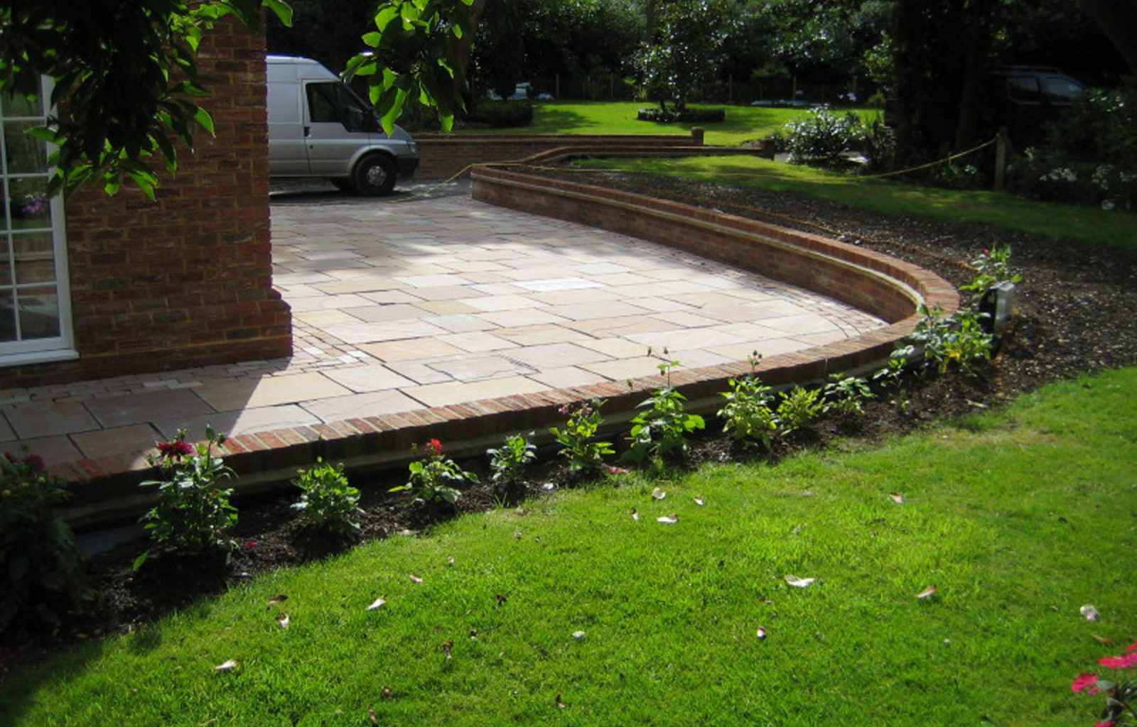 A large sunken patio area surrounding a brick house, with walled edging, lawn and planted border