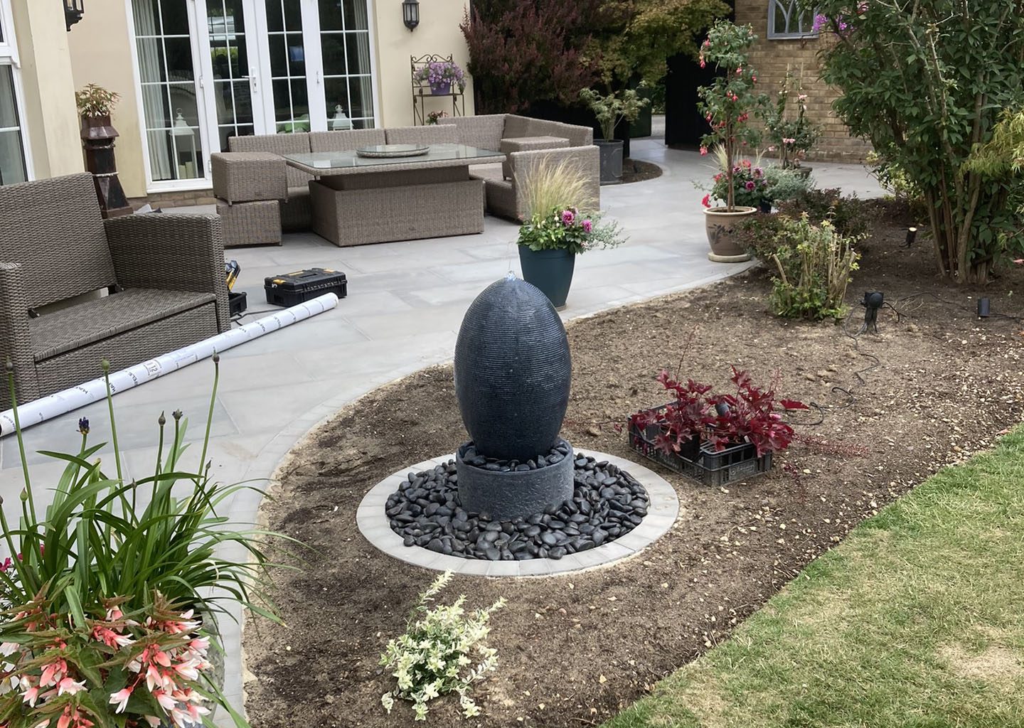 Sculpture water feature in plant border in front of paved patio area for garden seating