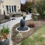 Sculpture water feature in plant border in front of paved patio area for garden seating