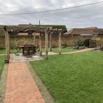 Landscaped garden enclosed by brick wall containing a wooden pergola with lighting in the middle of the garden over garden feature on paved area and brick herringbone pathway to and from area