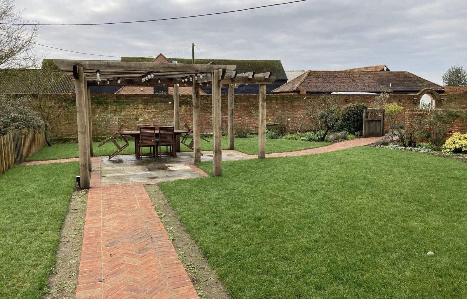 Landscaped garden enclosed by brick wall containing a wooden pergola with lighting in the middle of the garden over garden feature on paved area and brick herringbone pathway to and from area